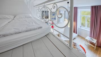 A bedroom with a bed, a window with curtains, a small table with a lamp, and a decorative metal railing.