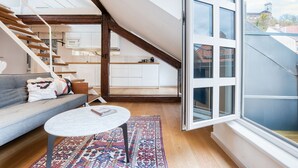 A modern loft apartment with a staircase leading to an upper floor, a kitchen area, and a large window offering a view of the city.
