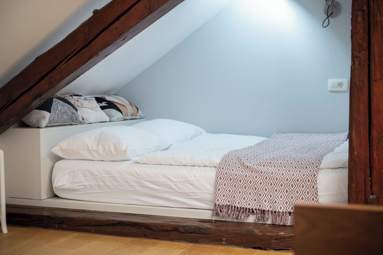 A bedroom with a sloped ceiling, a bed with white bedding, a patterned throw blanket, and a wooden beam structure.