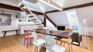A dining area with a wooden table, white chairs, and a chandelier. A staircase with wooden beams leads to an upper level.