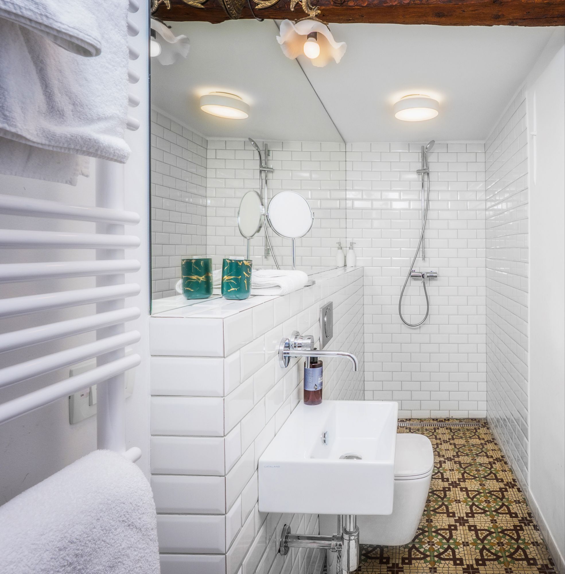 A modern bathroom with white tiles, a white sink, a toilet, and a shower. There are two round mirrors above the sink and a towel rack to the left.