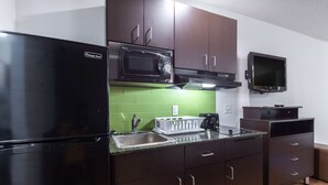 Cocina compacta con armarios oscuros, un refrigerador negro, un microondas, un lavamanos pequeño y un televisor montado sobre la encimera.