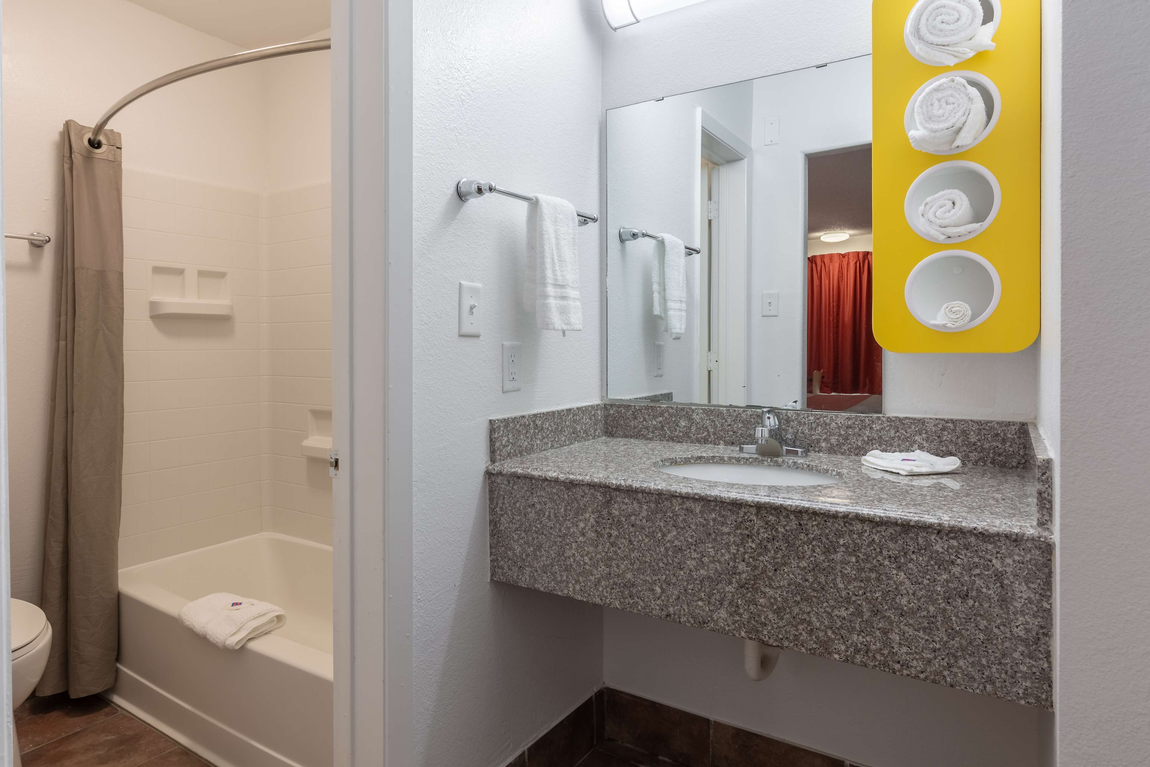 A bathroom with a shower, a sink with a granite countertop, and a mirror with a yellow towel holder.