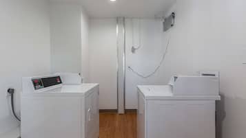 Un lavadero con dos lavadoras blancas, suelo de madera y una pared blanca con una rejilla de ventilación.
