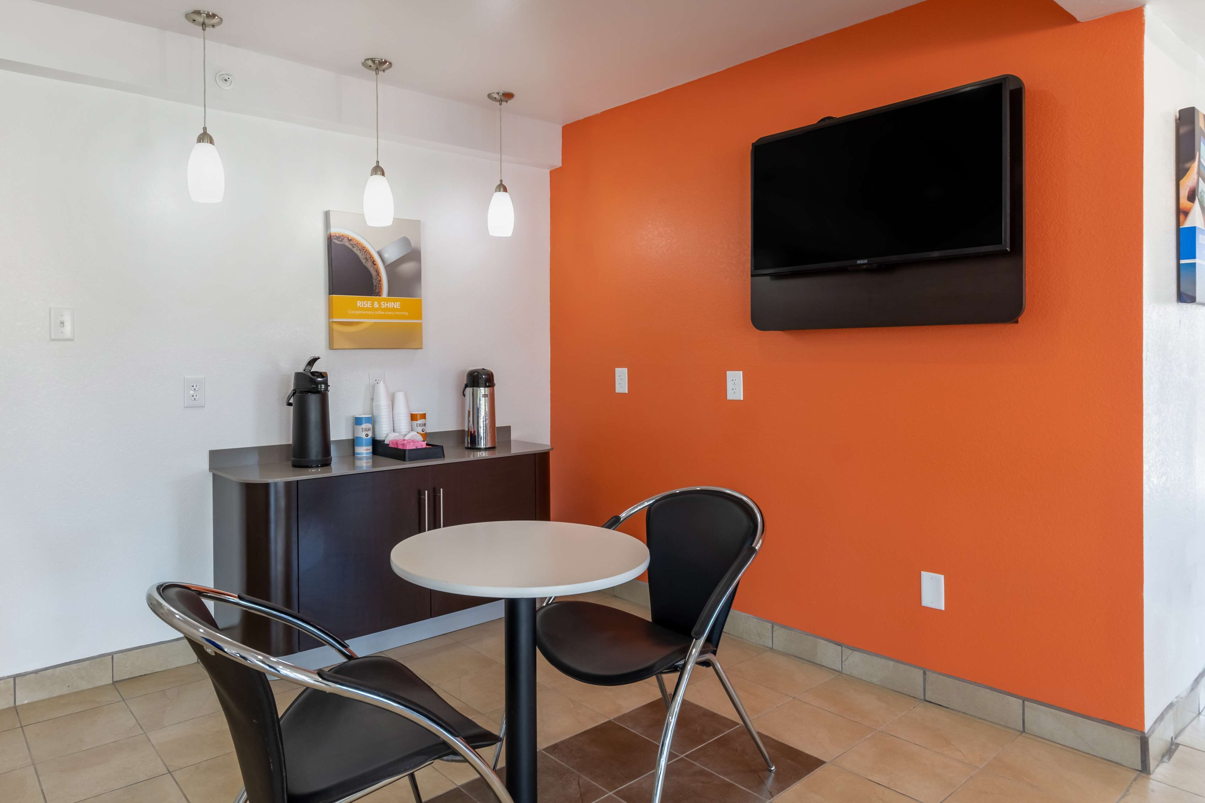 A small, modern room with a round table and two chairs, a flat-screen TV mounted on an orange wall, and a coffee machine on a dark cabinet.