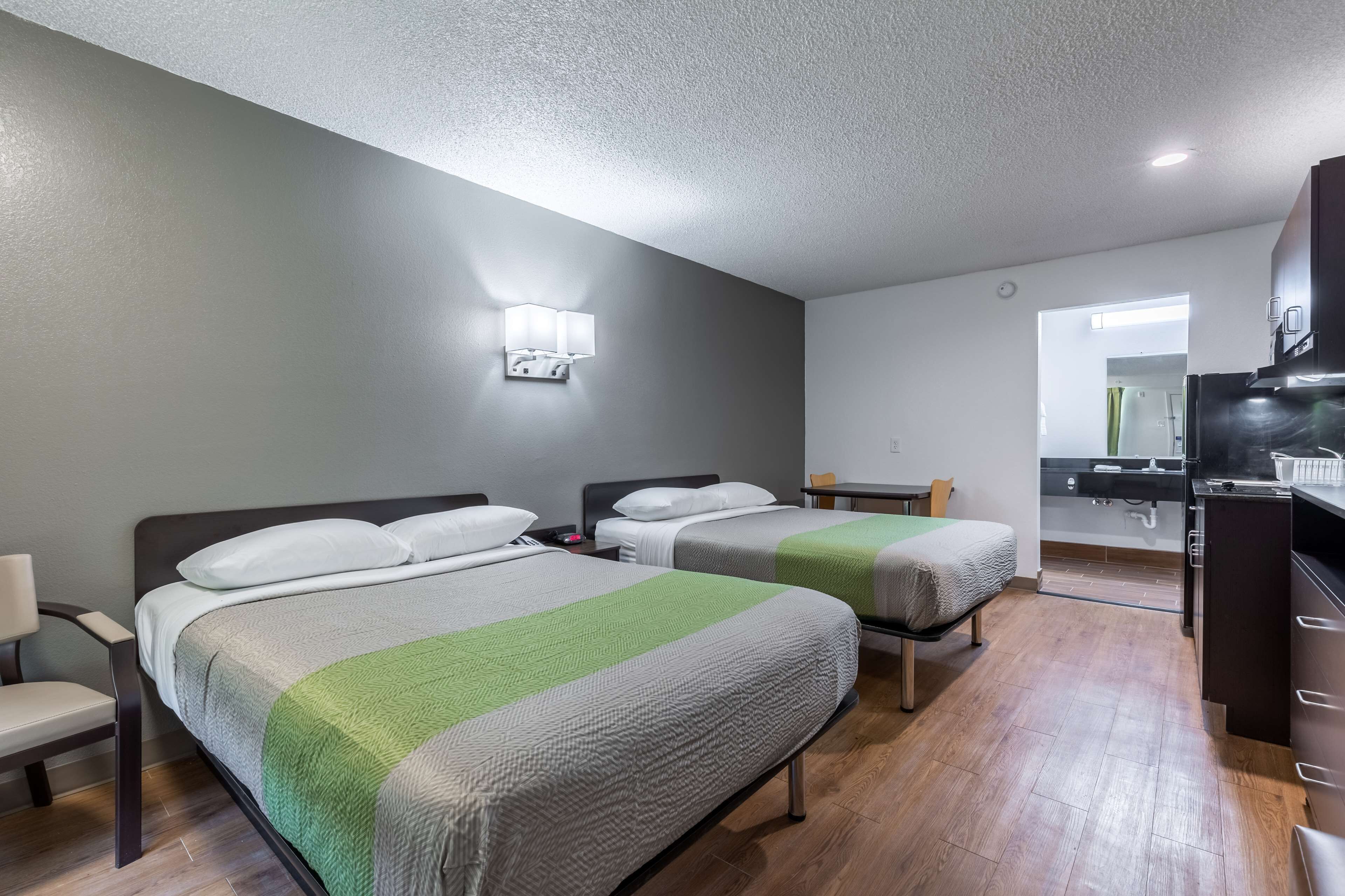 A hotel room with two beds, a desk, a chair, a small table, a mirror, and a kitchenette.
