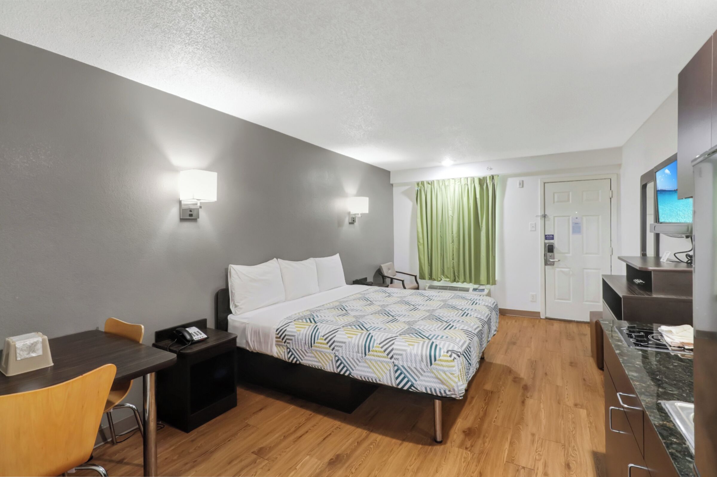 A hotel room with a bed, bedside table, chair, desk, and a small kitchenette.