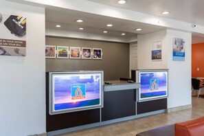 Un área de recepción con dos pantallas digitales grandes, un mostrador de recepción y una zona de estar con sillas naranjas.