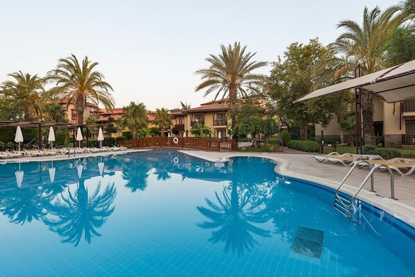 A swimming pool with lounge chairs and umbrellas, surrounded by palm trees and buildings. - Club Hotel Felicia Village - All Inclusive (Manavgat)