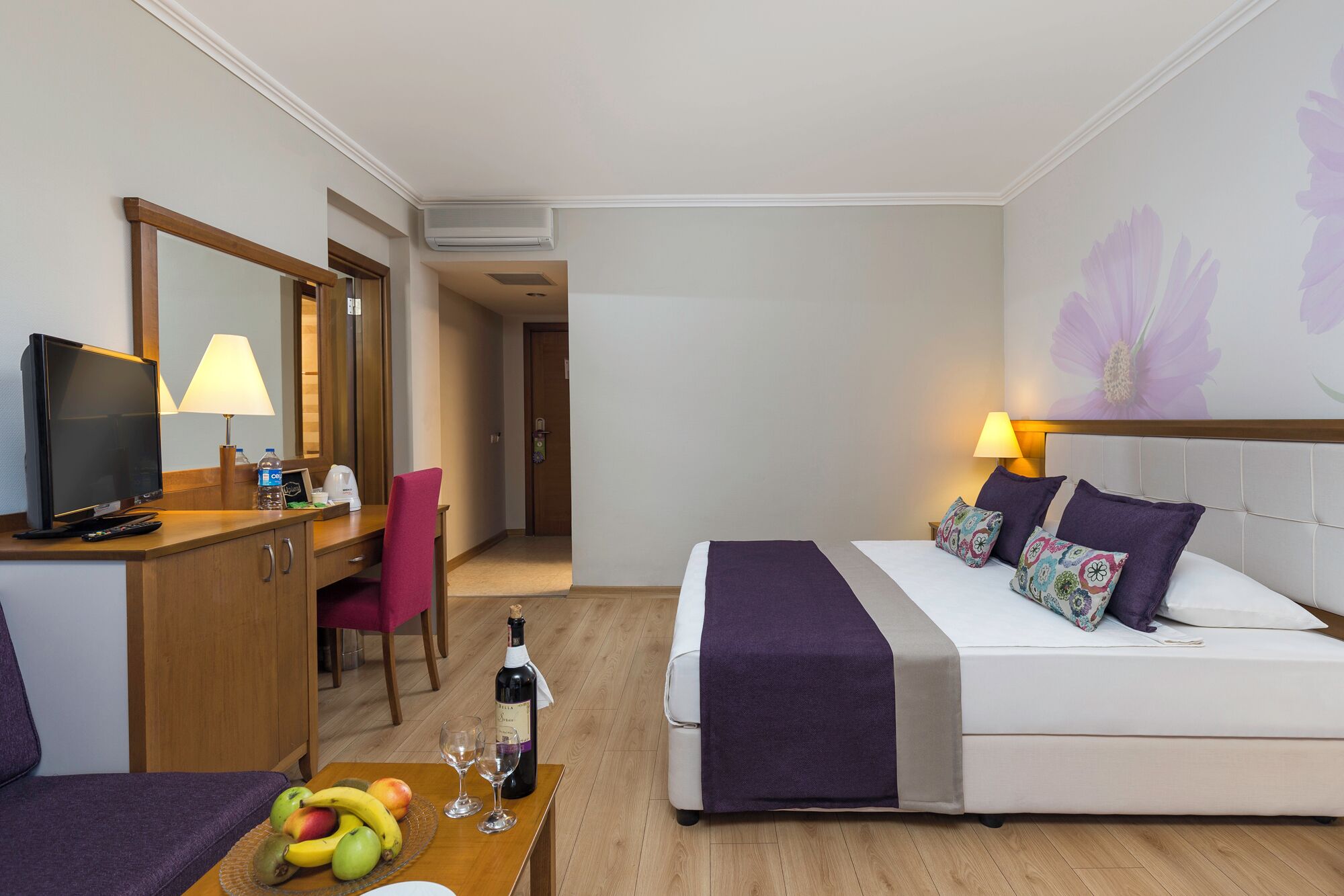 Ein Hotelzimmer mit Bett, Schreibtisch mit Fernseher, Sessel, Tisch mit Früchten und einer Weinflasche, sowie Wand mit floralem Dekor.