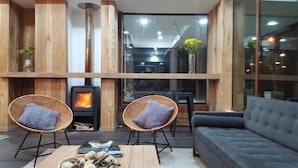 A cozy room with a wood-burning stove, two wicker chairs with cushions, a sofa, and a large window with a view of the city.