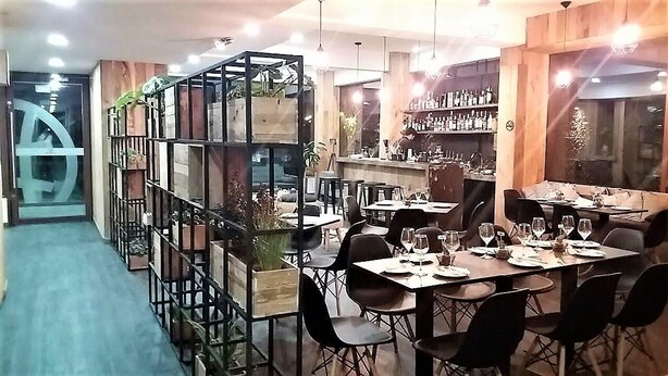A restaurant with multiple tables and chairs, a bar area with shelves of bottles, and a large window providing natural light.