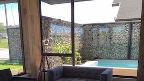 A modern living room with a sofa, a coffee table with books and a plant, and a large window with a view of a swimming pool and stone wall.
