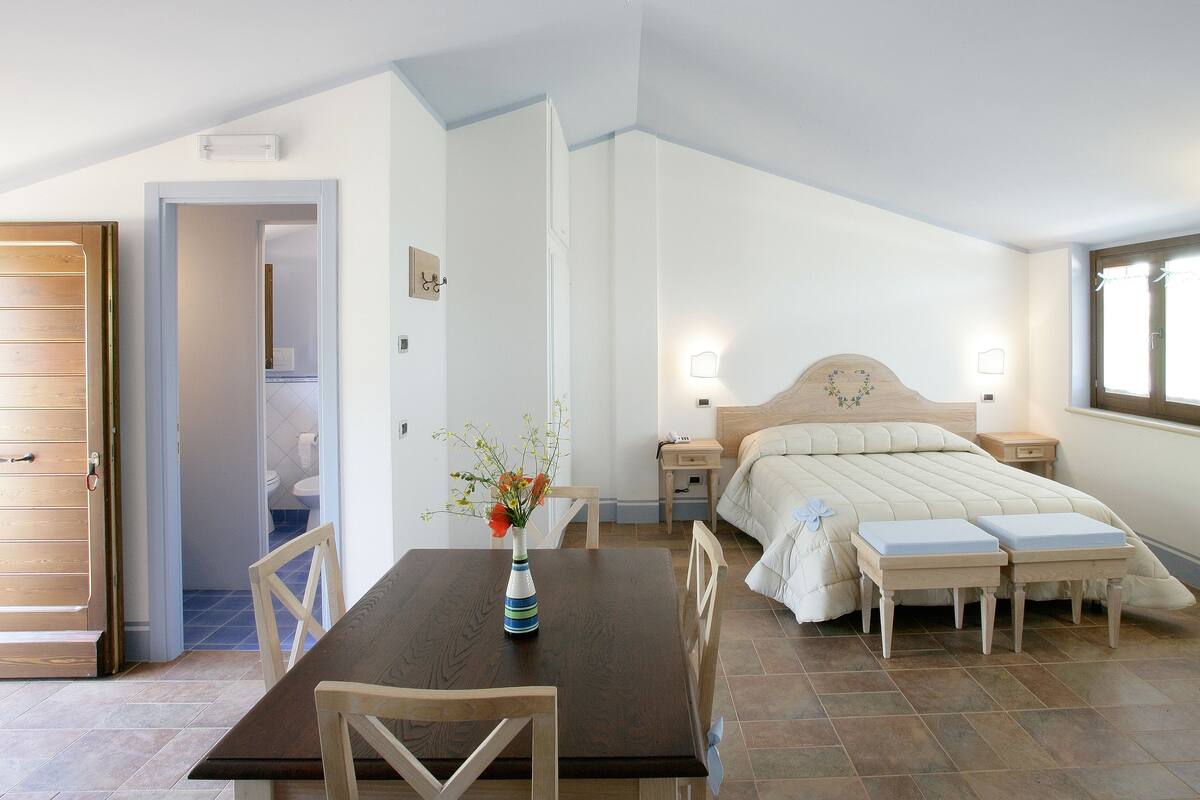 A bedroom with a bed, bedside tables, a nightstand, a wooden table with chairs, and a vase with flowers.