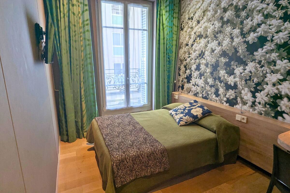 A bedroom with a bed, green curtains, a window with a view, and a floral wallpaper.