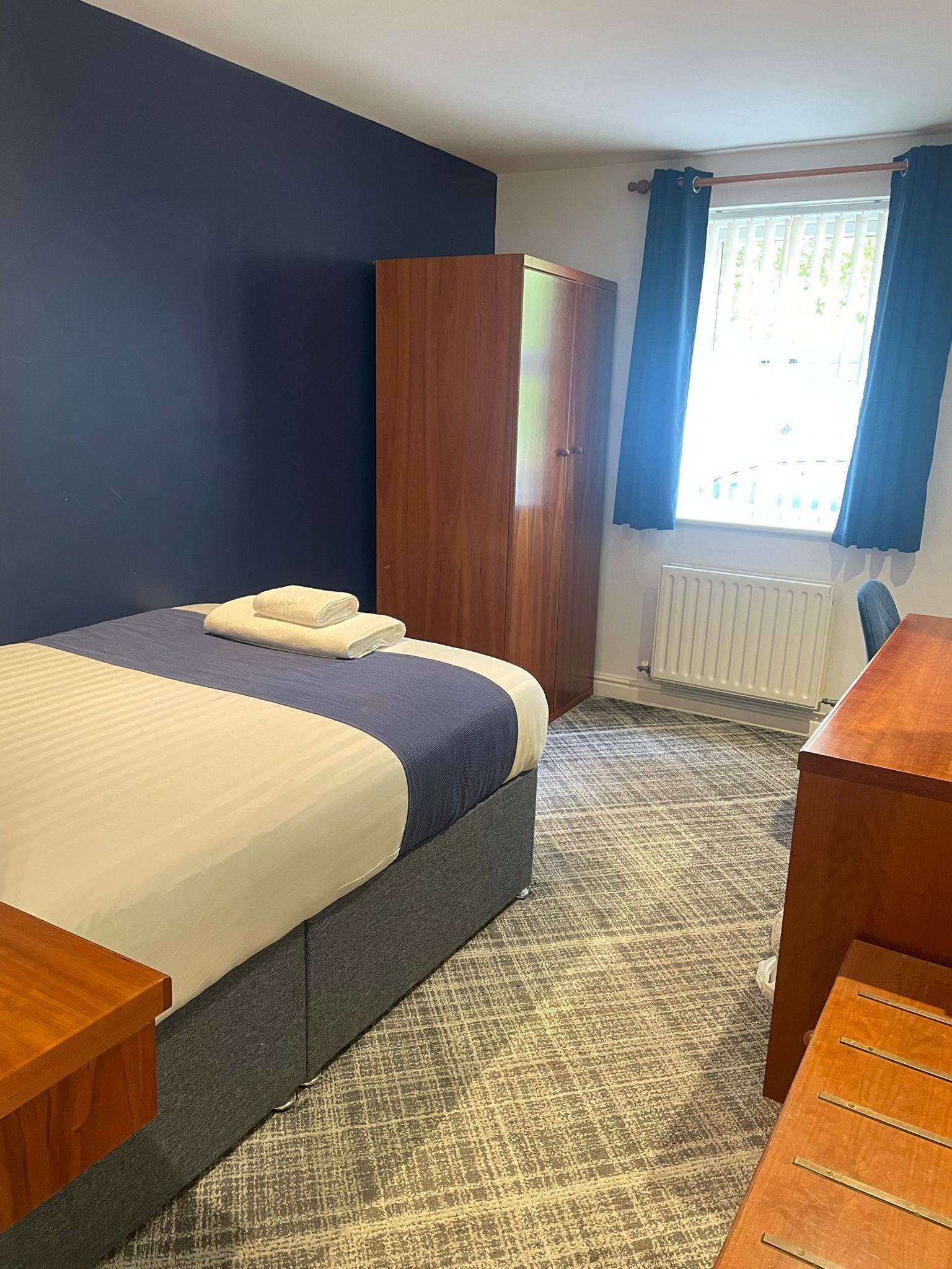 A hotel room with a bed, a wooden wardrobe, a desk, and a window with curtains.