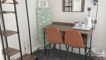 Una habitación pequeña con una mesa de madera, dos sillas naranjas, una tabla de planchar blanca, un espejo y una cafetera.