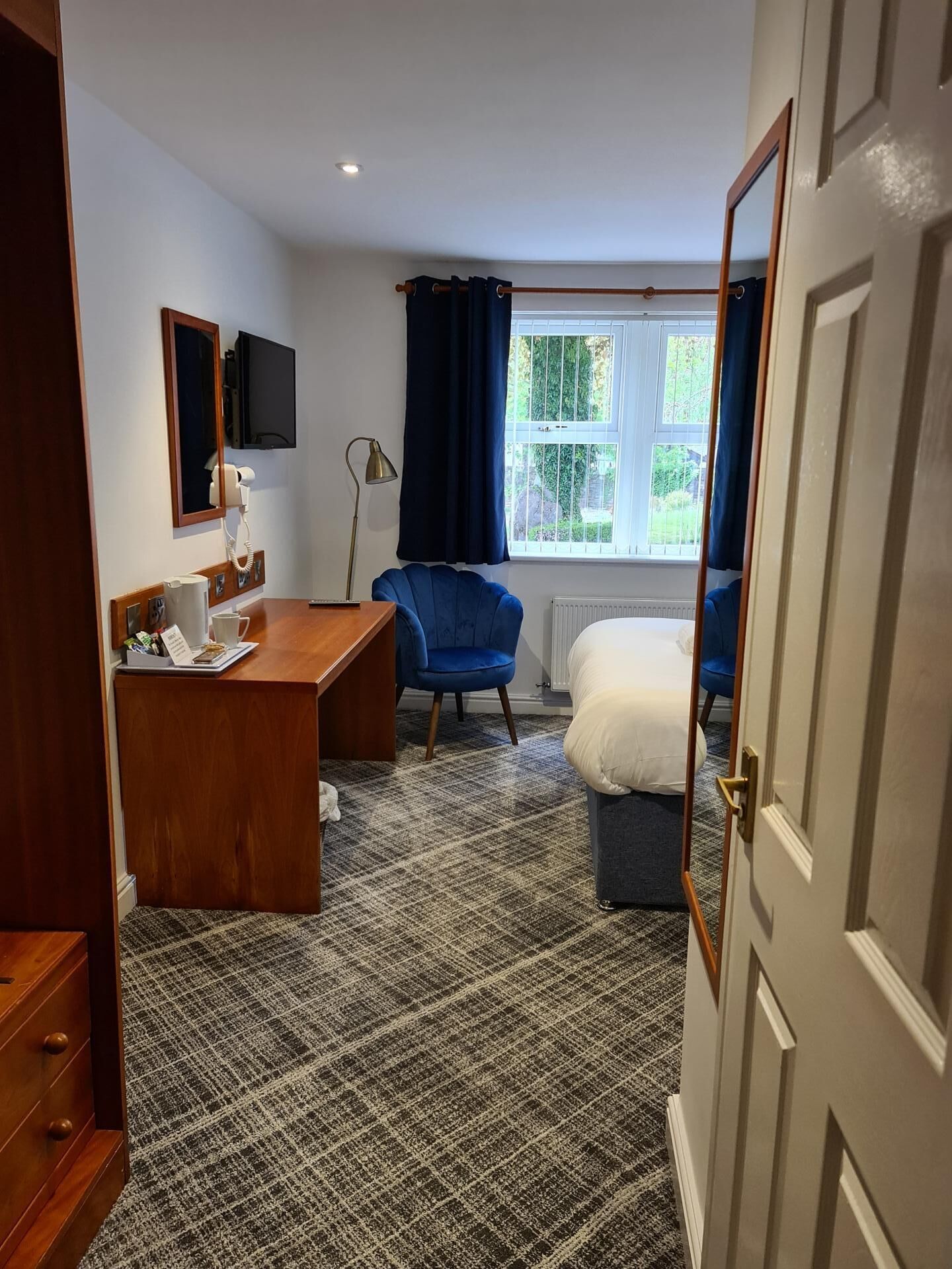 Habitación de hotel con un escritorio de madera, un sillón azul, una cama, un televisor de pantalla plana y una ventana con cortinas.