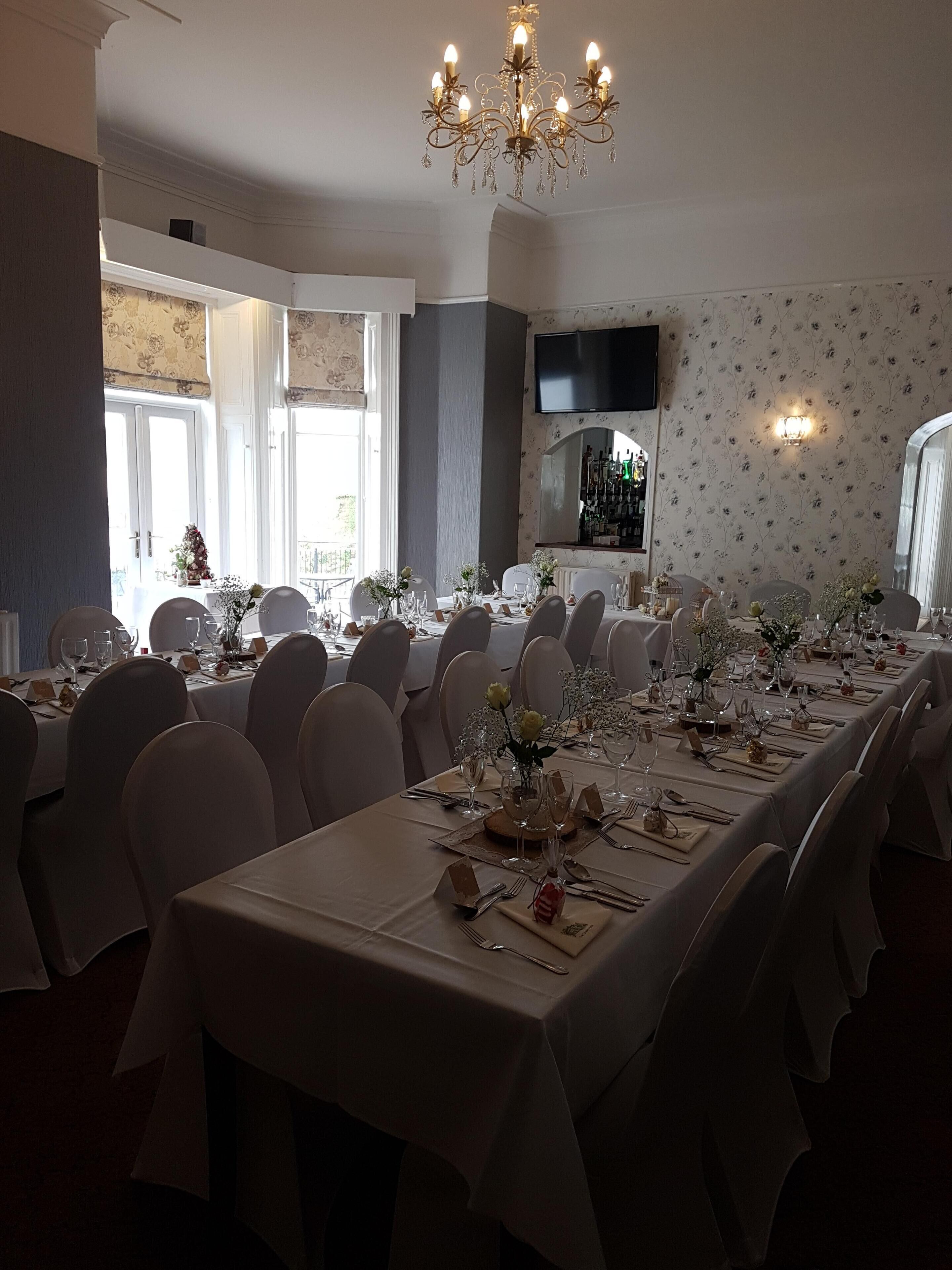 A formal dining room with long tables set for a meal, a chandelier, and a wine bar in the background.