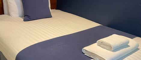 Una cama bien hecha con ropa de cama blanca y azul, una toalla blanca doblada y un cabecero de madera.