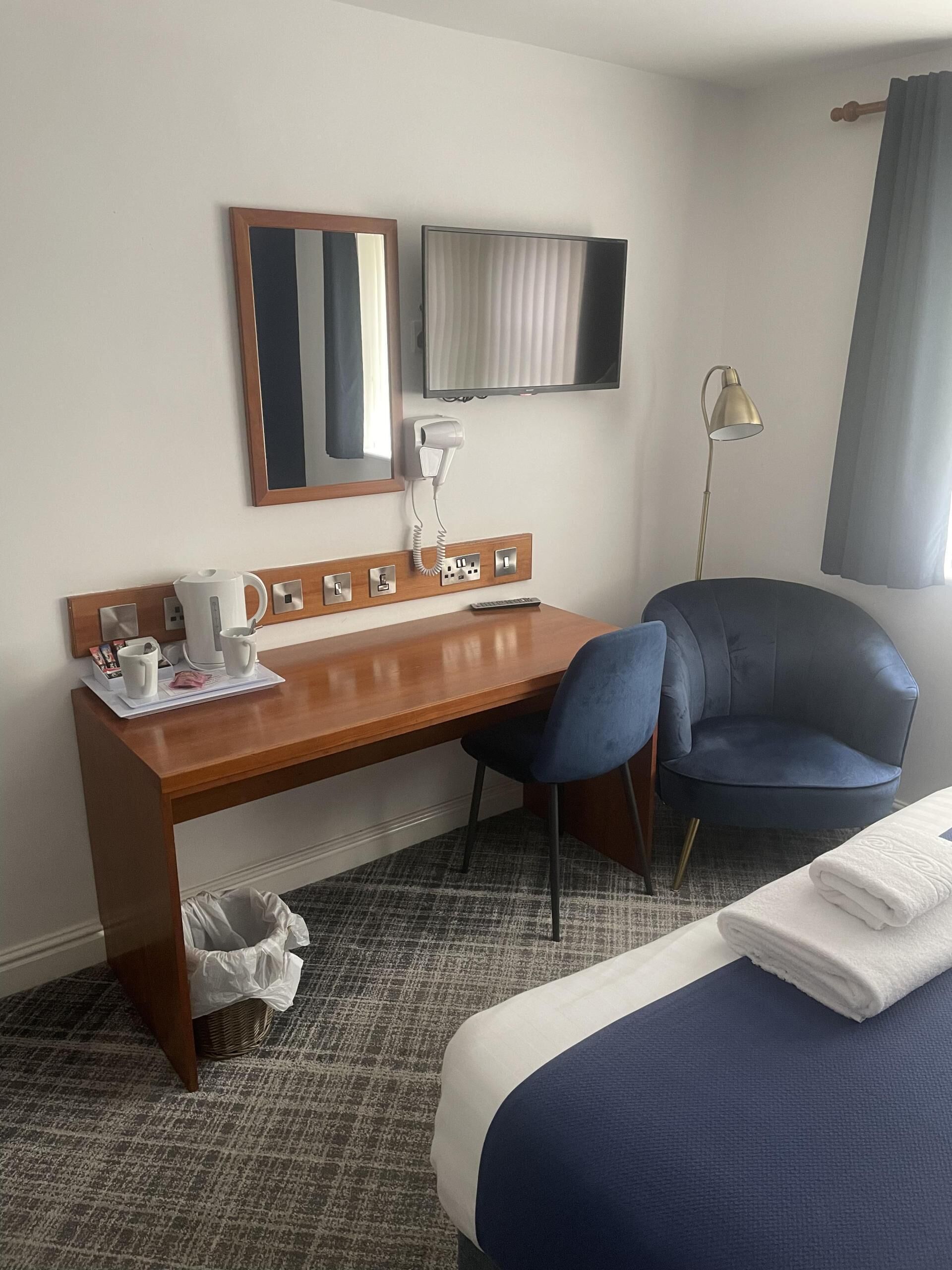 A hotel room with a wooden desk, a chair, a bed, a mirror, a TV, a lamp, and a window with curtains.