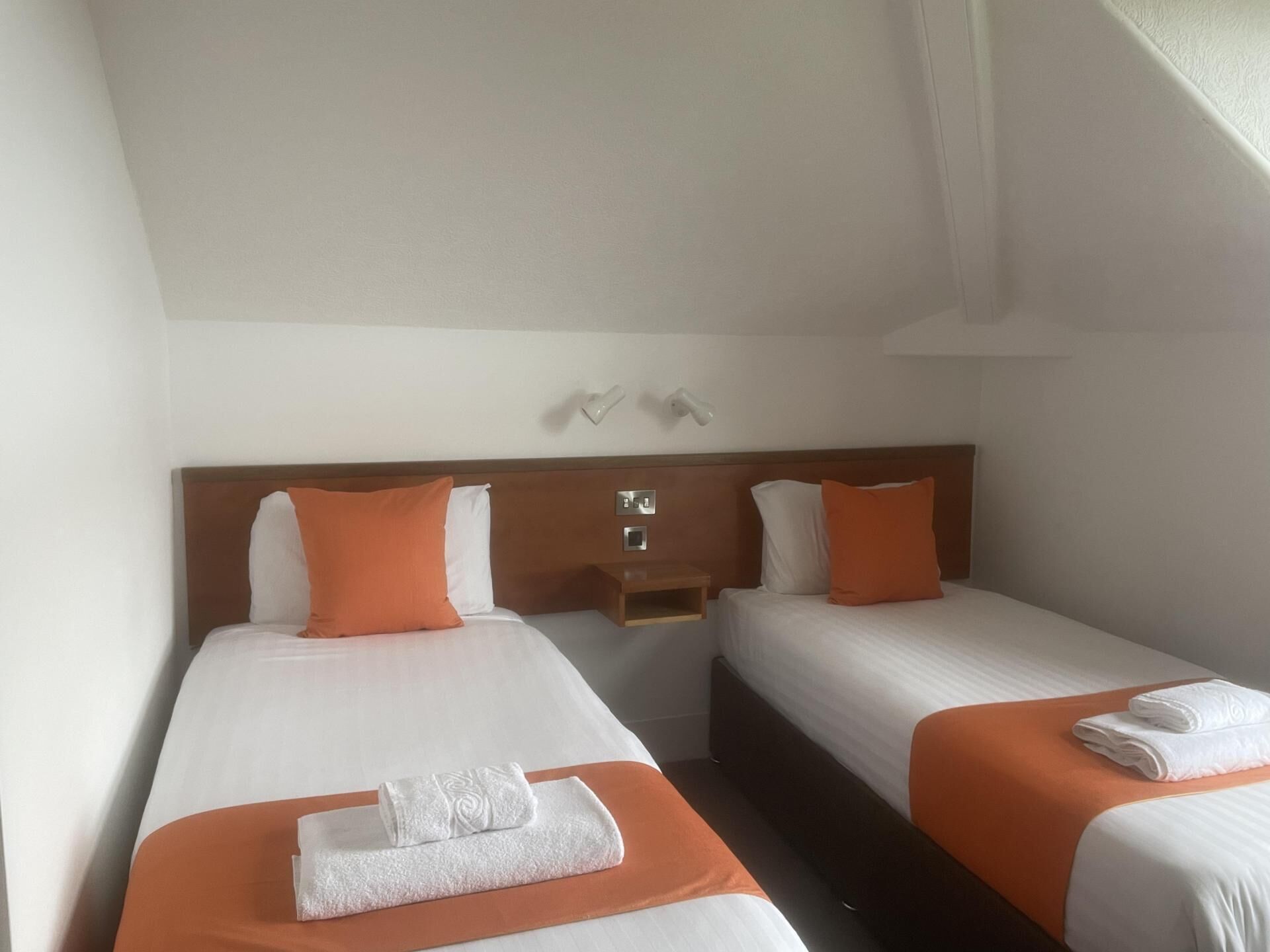 Habitación de hotel con dos camas, cada una con una colcha naranja y almohadas blancas. Hay dos mesitas de noche con lámparas y un pequeño estante sobre las camas.
