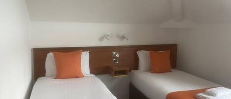 Habitación de hotel con dos camas, cada una con una colcha naranja y almohadas blancas. Hay dos mesitas de noche con lámparas y un pequeño estante sobre las camas.