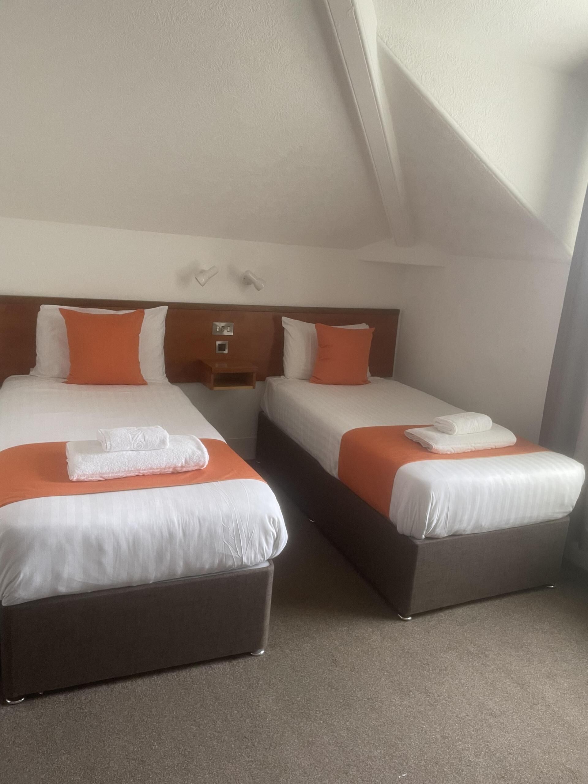 Habitación de hotel con dos camas, cada una con ropa de cama blanca y almohadas naranjas. Hay dos mesitas de noche con lámparas, y una pequeña mesita de noche de madera con una lámpara. La habitación tiene un techo inclinado y suelo alfombrado.