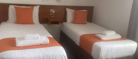 Habitación de hotel con dos camas, cada una con ropa de cama blanca y almohadas naranjas. Hay dos mesitas de noche con lámparas, y una pequeña mesita de noche de madera con una lámpara. La habitación tiene un techo inclinado y suelo alfombrado.