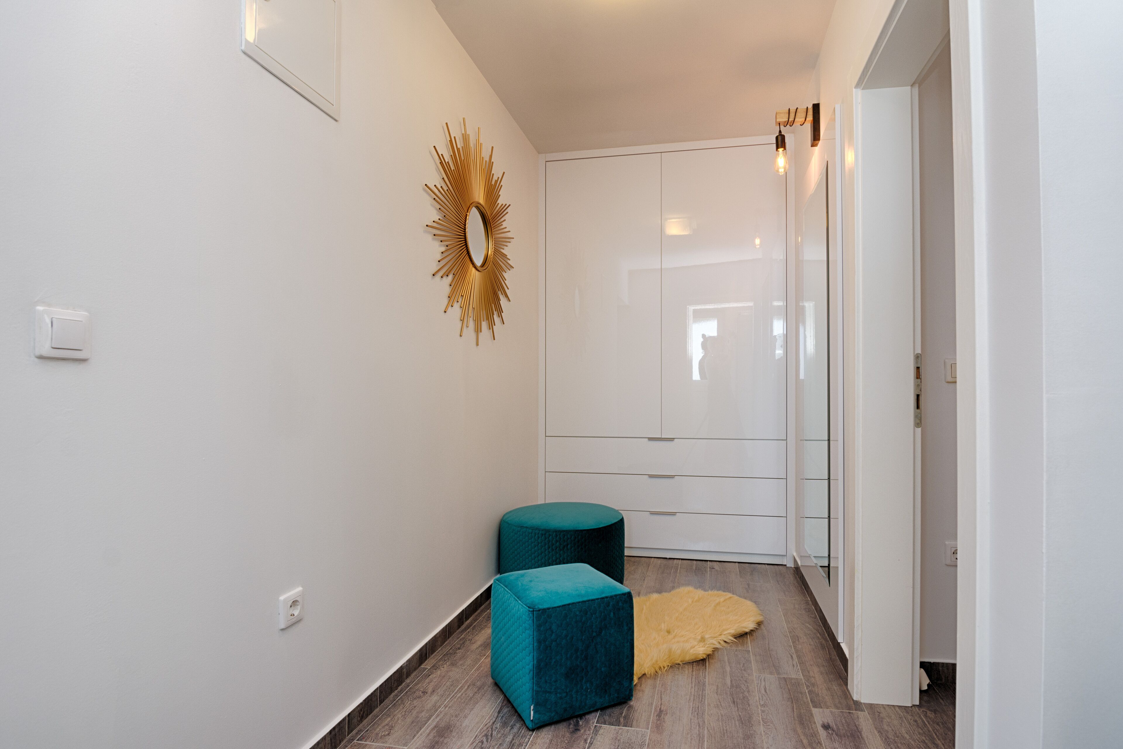 A hallway with white walls, a wooden floor, a teal ottoman, a gold mirror, and a door with a glass panel.