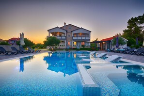 A swimming pool with lounge chairs and umbrellas, surrounded by a building with balconies and a red roof. - Apartments Basic (Vir)