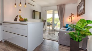 A modern kitchen with a central island, a dining area with a grey sofa and blue cushions, and a large window with a view of greenery. - Apartments Basic (Vir)