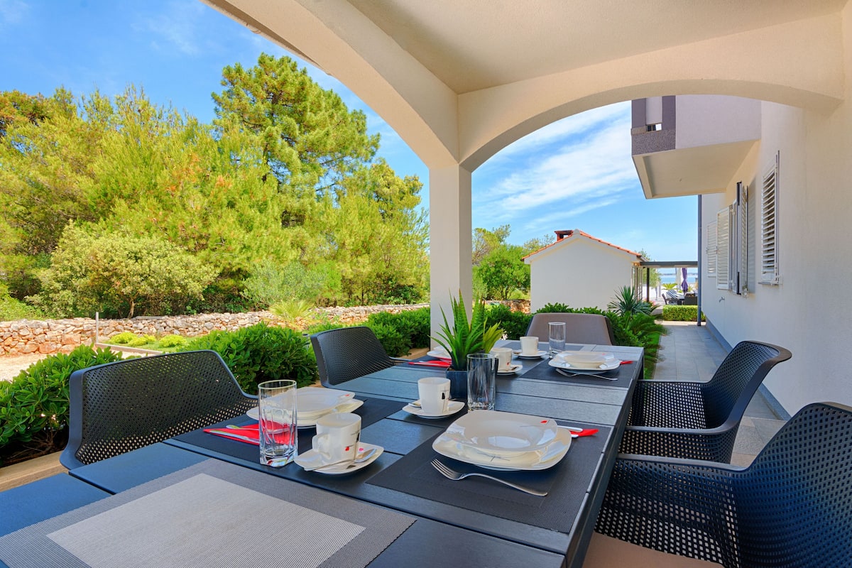 An outdoor dining area with a table set for four, surrounded by chairs, and overlooking a garden with trees and a stone wall.