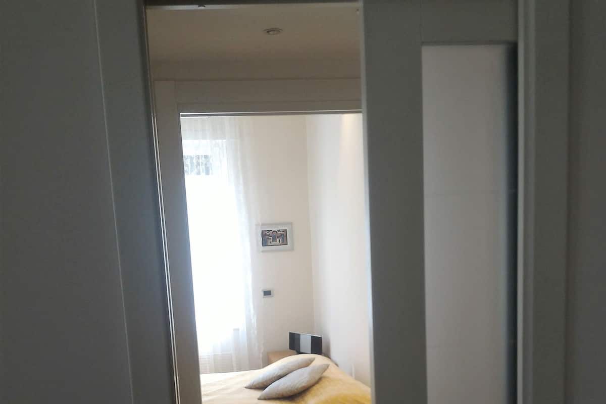 A bedroom with a bed, wooden flooring, and a door leading to another room.