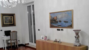 Habitación con piso de madera, un candelabro, un cuadro de una escena costera, un aparador con objetos decorativos y una silla.