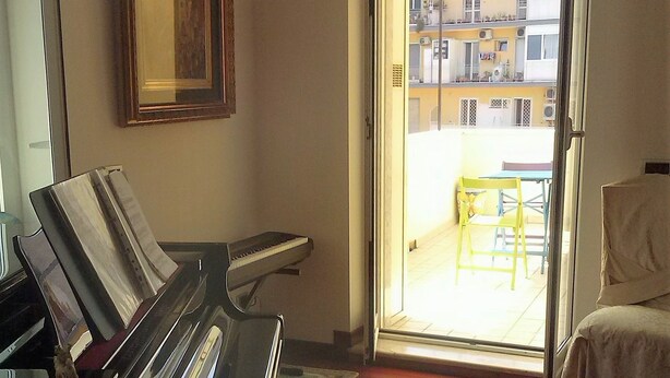 Una habitación con un piano, una pequeña mesa y una silla. Hay una puerta que da a un espacio exterior con vistas a edificios.