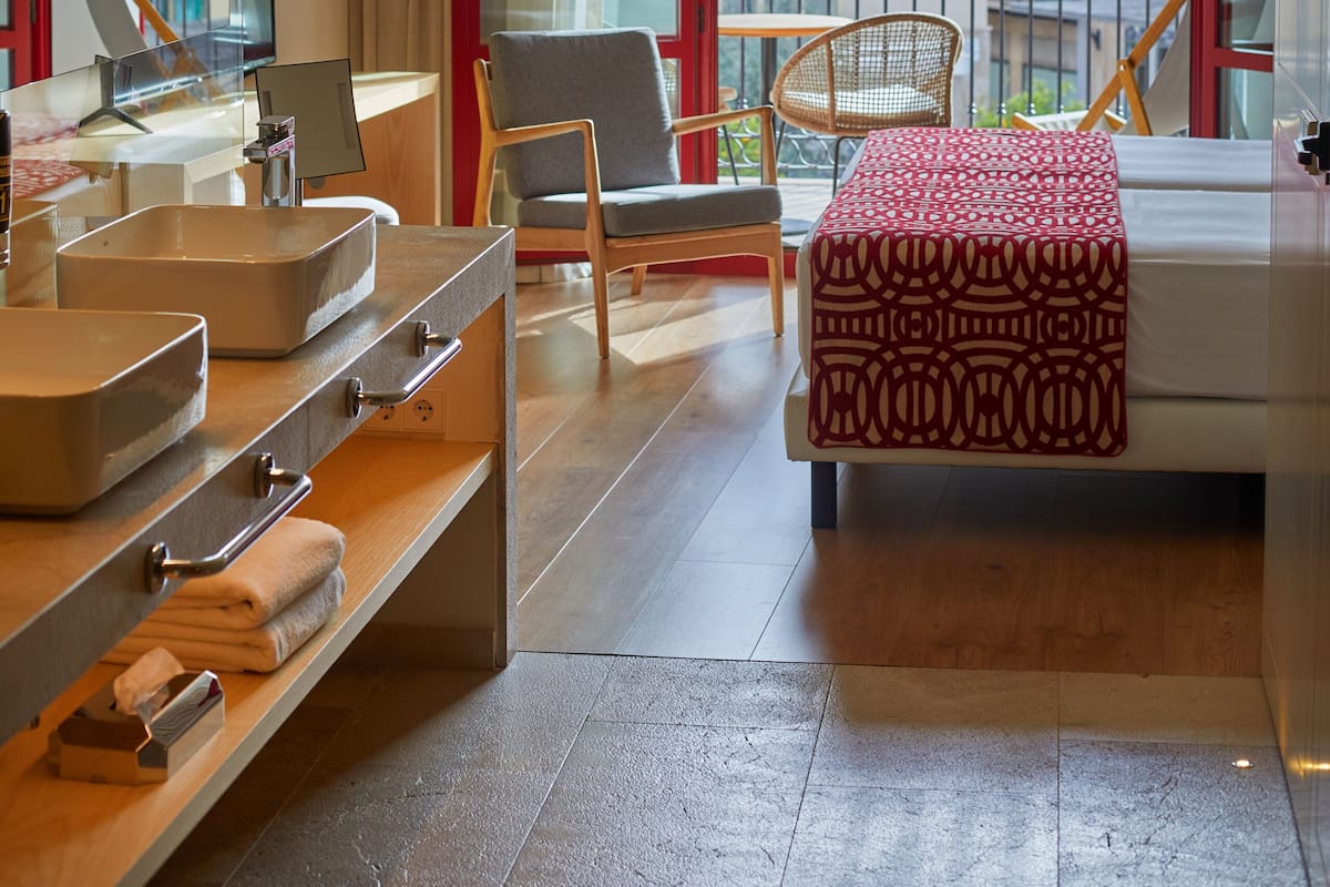 Ein Hotelzimmer mit großem Fenster, einem Holztisch, einem Stuhl, einem Bett mit gemustertem Überwurf und einem kleinen Tisch mit Laptop und Tasse.