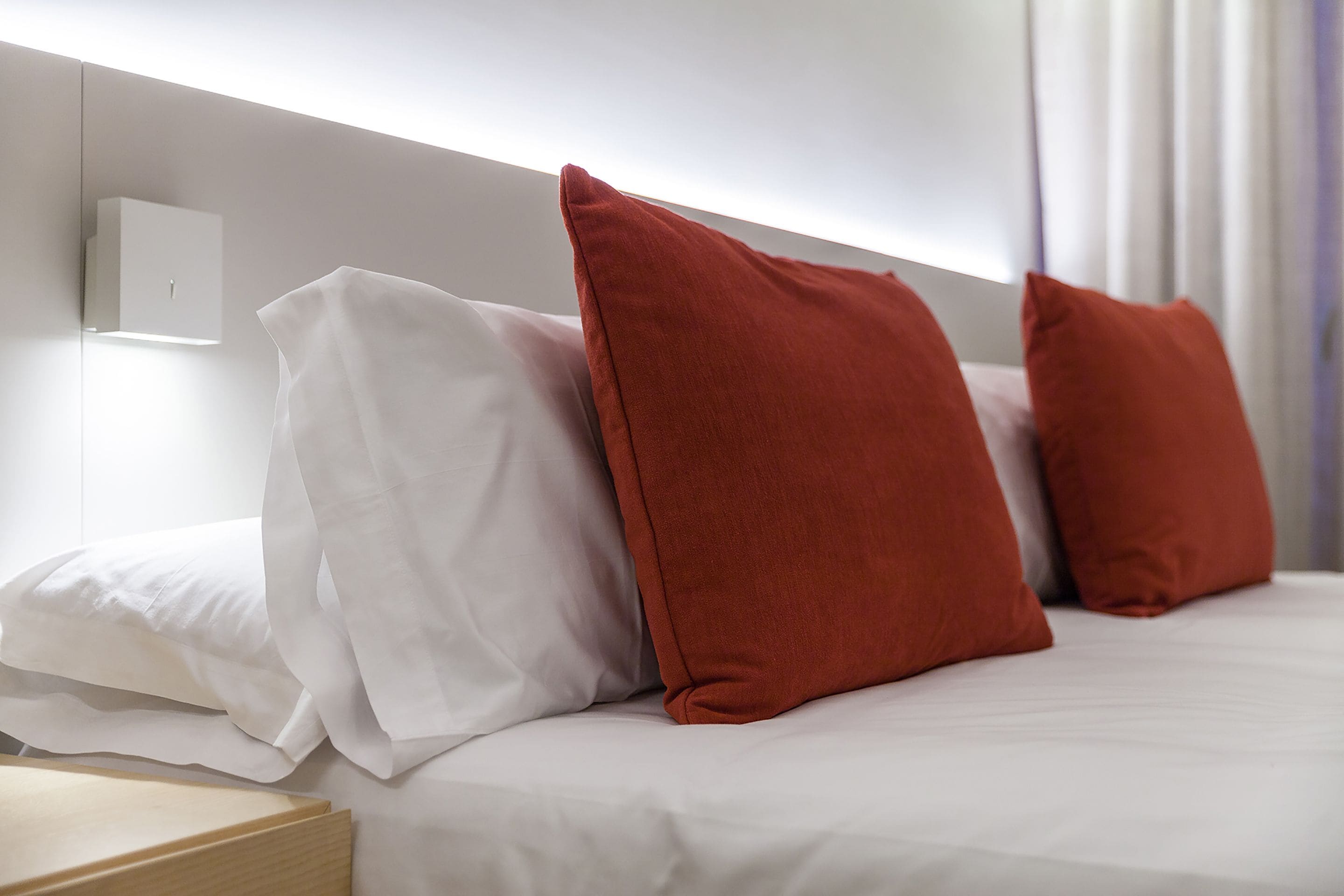 A neatly made bed with white and red pillows, a wooden bedside table, and a white wall with a light switch.