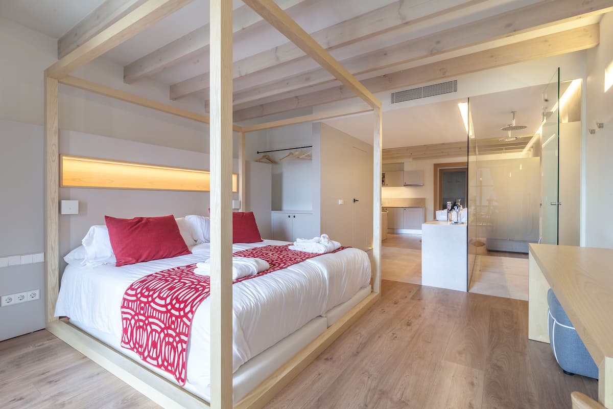 Ein modernes Schlafzimmer mit einem großen Bett, Holzbalken, einem Nachttisch und einem im Hintergrund sichtbaren Badezimmer.