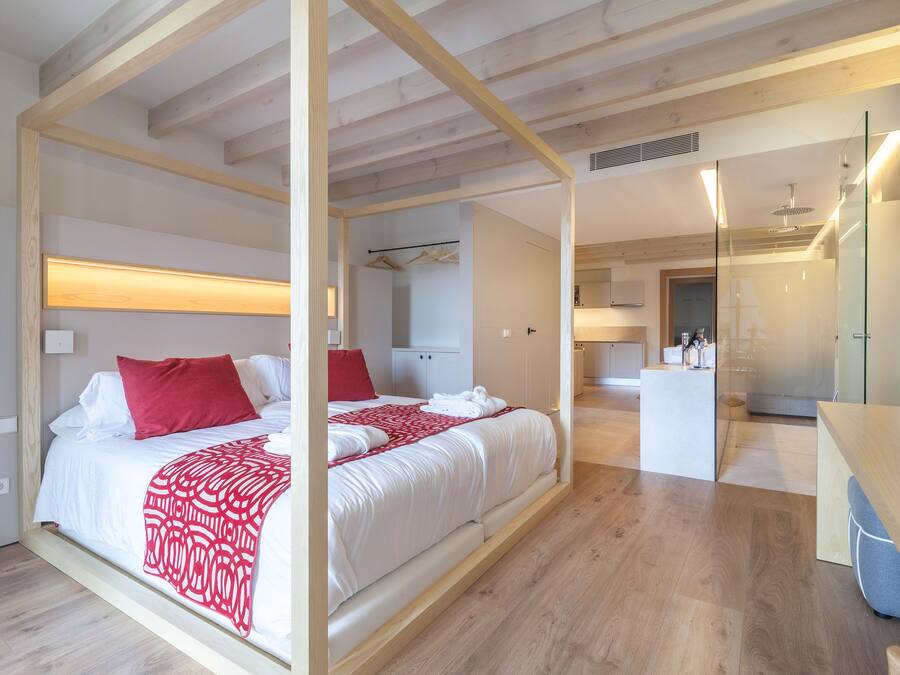 A modern bedroom with a large bed, wooden beams, a bedside table, and a bathroom visible in the background.