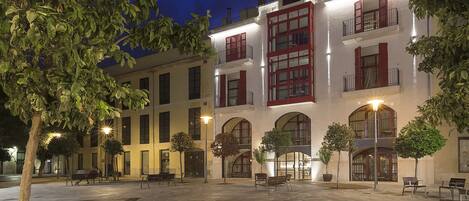 A courtyard with benches and potted plants, surrounded by buildings with red shutters and arched windows.