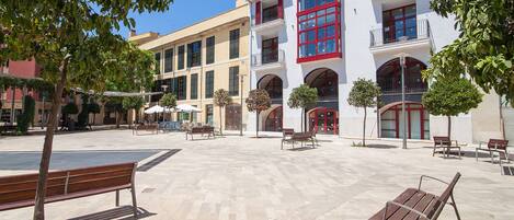 Un patio con bancos, árboles y un edificio de paredes blancas y contraventanas rojas.