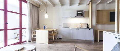 A modern kitchen with white cabinets, a microwave, and a coffee machine. There is a dining area with a table and chairs, and a large window with curtains.