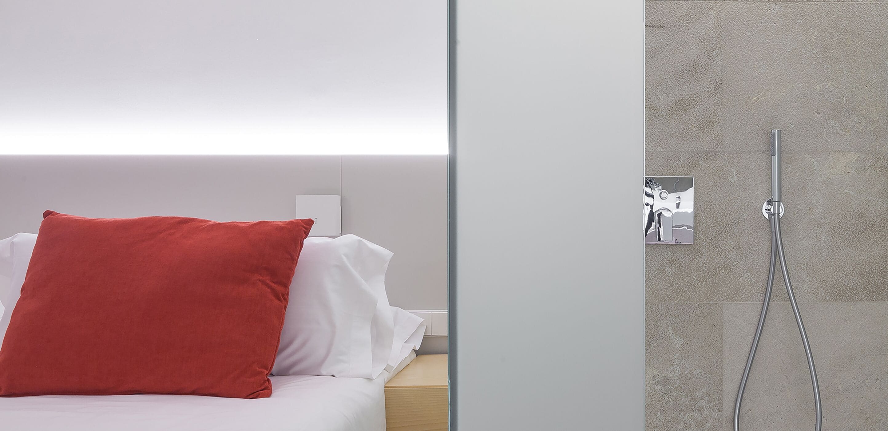 A modern hotel room with a bed, a red pillow, a white pillow, and a shower with a metallic handle.