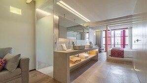 A modern bathroom with a glass partition, a vanity with a mirror, and a bed with a red and white patterned blanket. -  Apartahotel Fil Suites (Palma de Mallorca)
