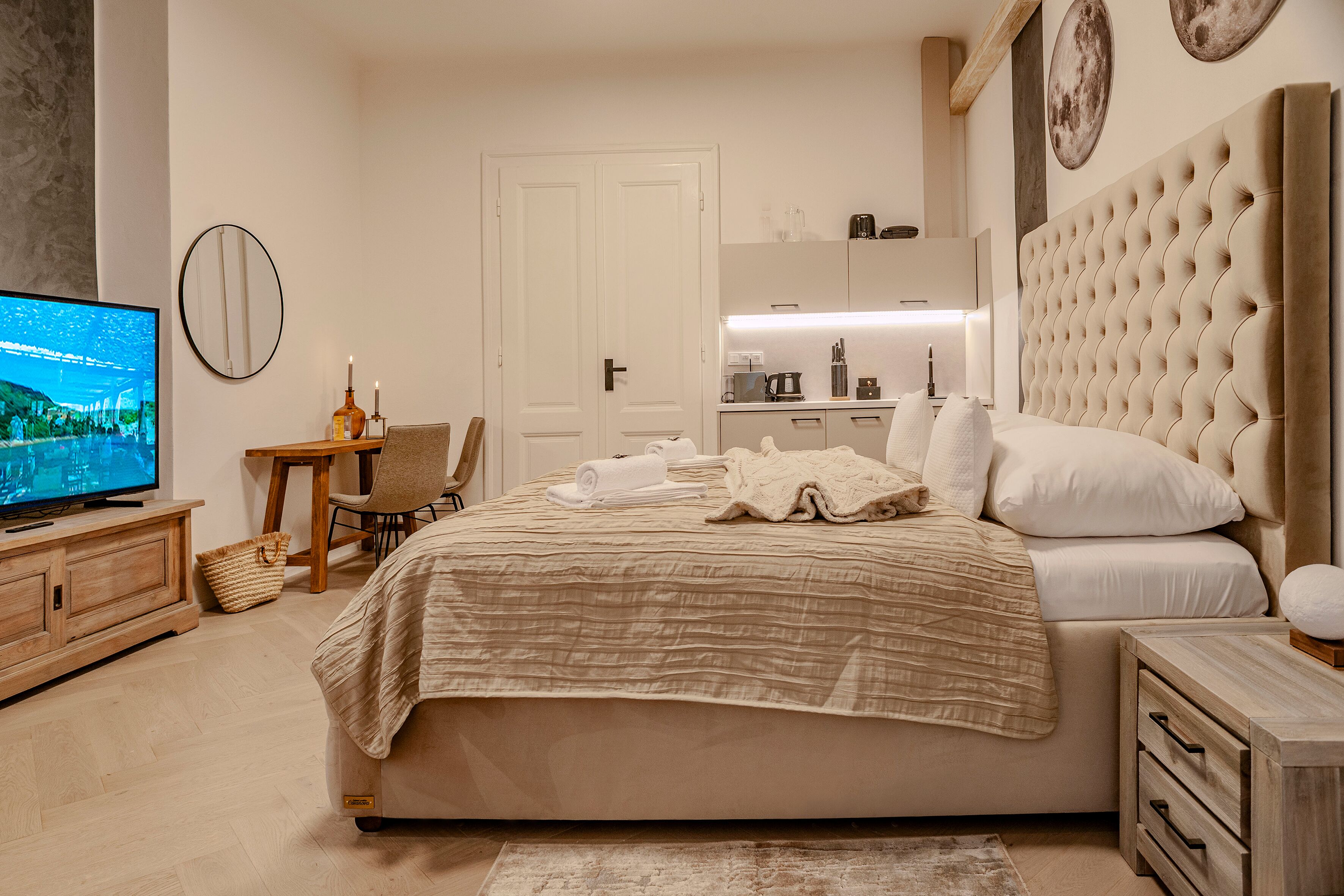 A bedroom with a large bed, a television, a wooden dresser, and a kitchen area in the background.