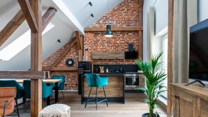Un espacio habitable tipo loft con vigas de madera a la vista, pared de ladrillo, área de cocina con electrodomésticos modernos, sillas color turquesa y un televisor grande.