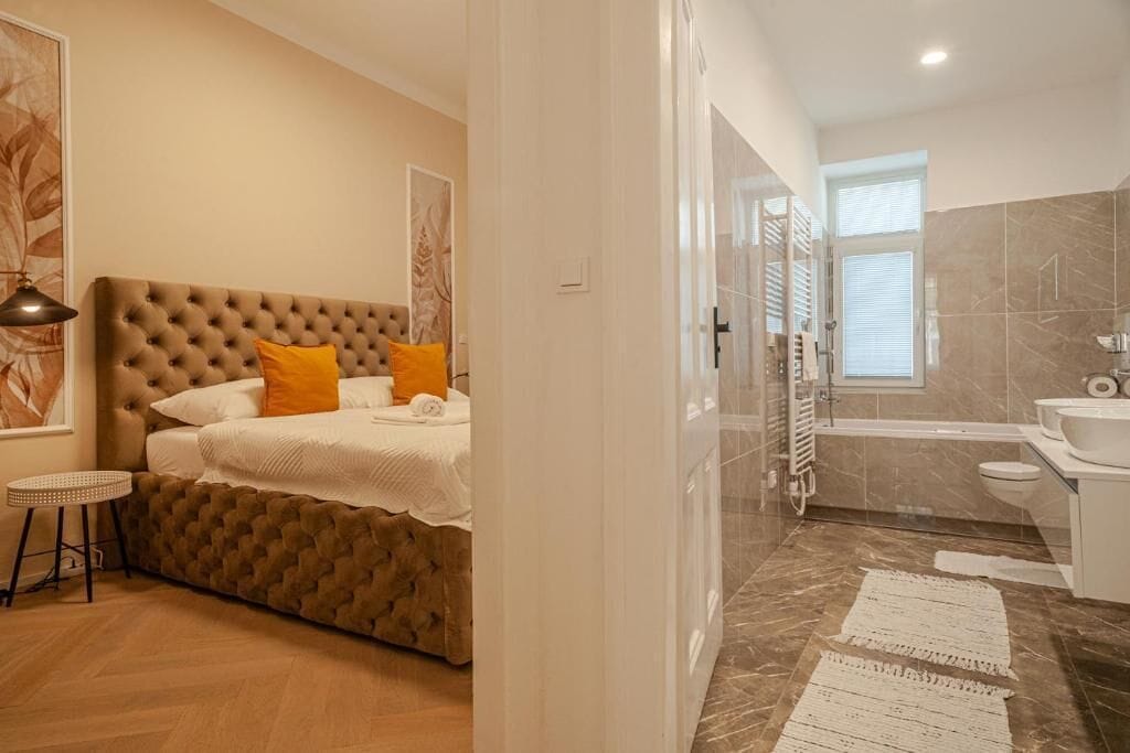 Un dormitorio con cabecera capitonada, cama con ropa de cama blanca y almohadas naranjas, una mesita redonda pequeña y un ventanal grande.