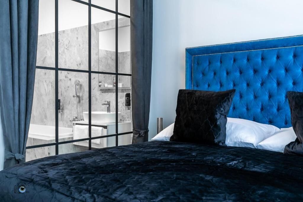 Ein Schlafzimmer mit großem Fenster, einem blauen, gepolsterten Kopfteil und Blick auf ein Badezimmer.
