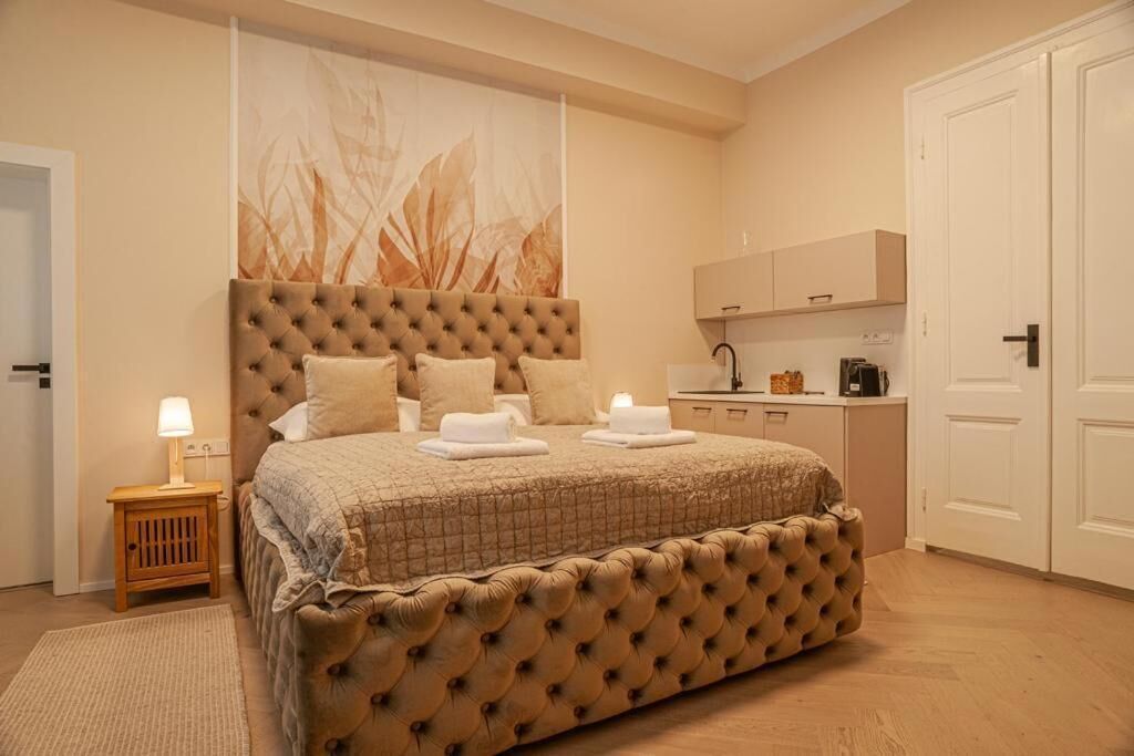 A bedroom with a large bed, a bedside table with a lamp, a wall-mounted artwork, a kitchenette, and a door.